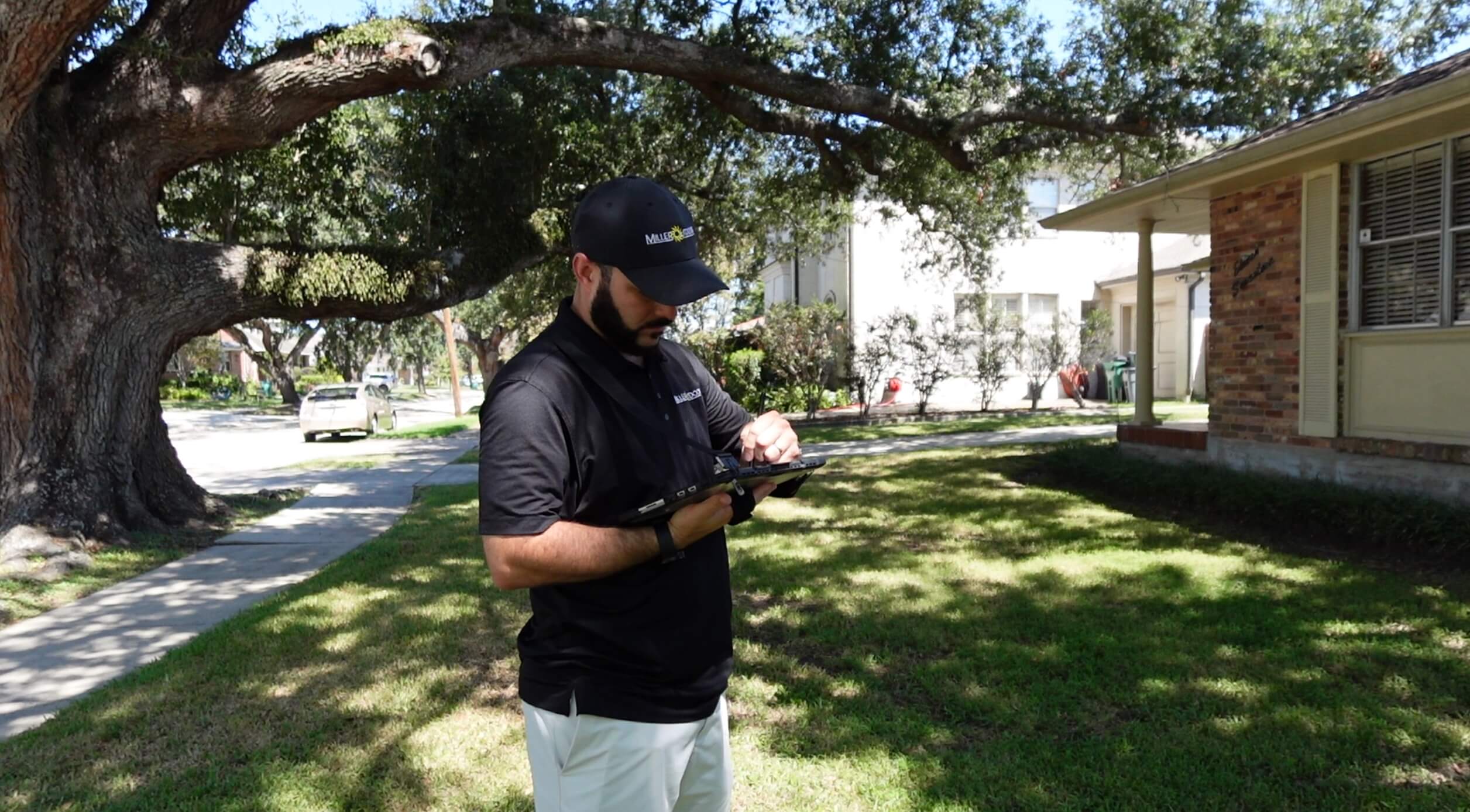 A man wearing a black cap and shirt stands on a grassy lawn with improved landscaping under a large tree, using a tablet. Houses and a sidewalk are visible in the background on a sunny day.