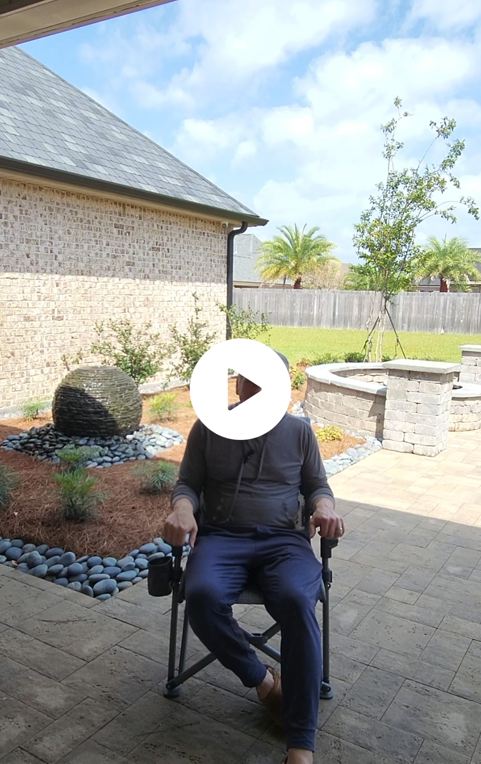A person sits on a chair in a sunny backyard patio with landscaped gardens, showcasing expert Landscape Development in New Orleans, featuring a circular stone fire pit, river rocks, a brick wall, and a distant palm tree.