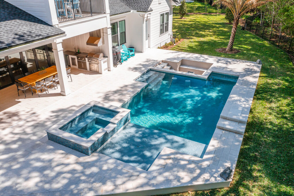 A backyard with a rectangular swimming pool, attached hot tub, and built-in outdoor kitchen and dining area—showcasing expert Pool Installation New Orleans—surrounded by a spacious tiled patio and lawn with trees.