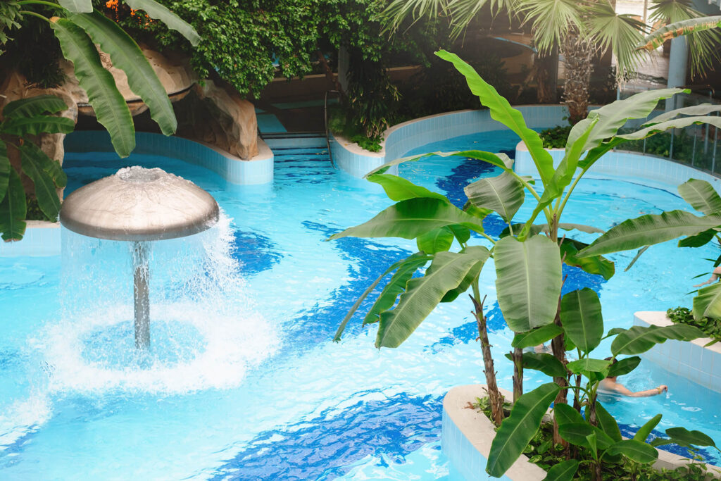 A bright blue indoor swimming pool with a mushroom-shaped water fountain and lush tropical plants, including banana trees, surrounding the pool area. Palm trees and greenery create a relaxing atmosphere, perfect for Pool Installation New Orleans style.