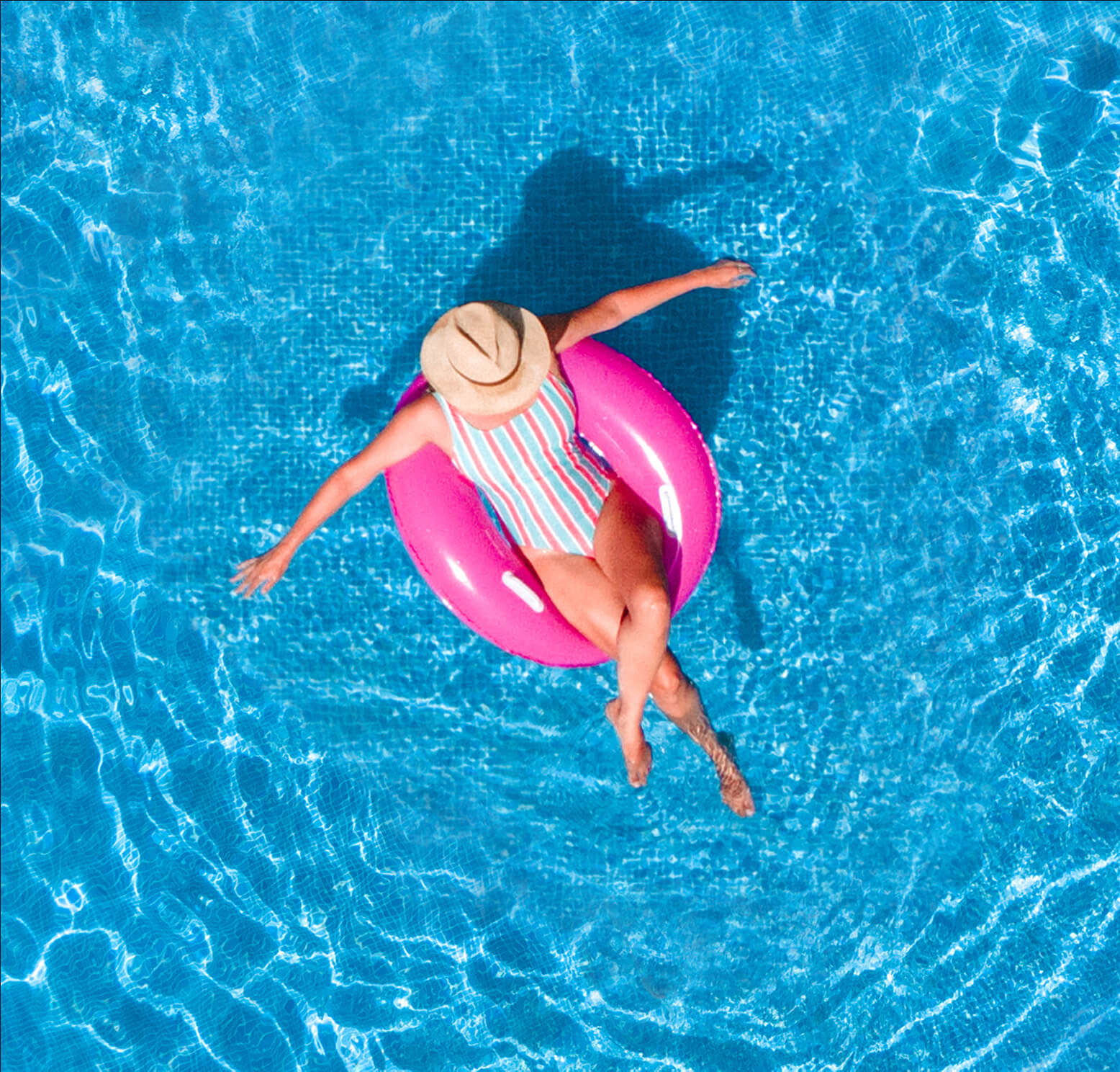 A person in a striped swimsuit and straw hat relaxes on a pink inflatable ring, floating in a clear blue swimming pool—an inviting scene that showcases the results of expert pool installation in New Orleans, viewed from above.
