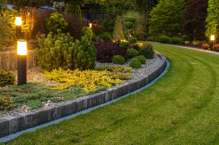A well-lit, landscaped garden with a curved stone border, various shrubs, small bushes, and outdoor lamp posts lining a neatly mowed premium lawn in the evening.