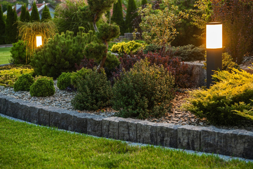 A landscaped garden with various shrubs and plants, bordered by a stone edge and illuminated by two modern outdoor solar landscape lights glowing warmly at dusk—an energy-efficient way to save money on your lighting.