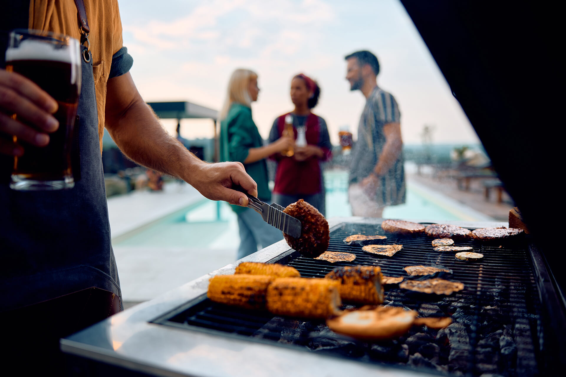 Person grilling corn, burgers, and vegetables on a barbecue by a pool, holding a drink, while three people stand in the background talking and socializing outdoors.