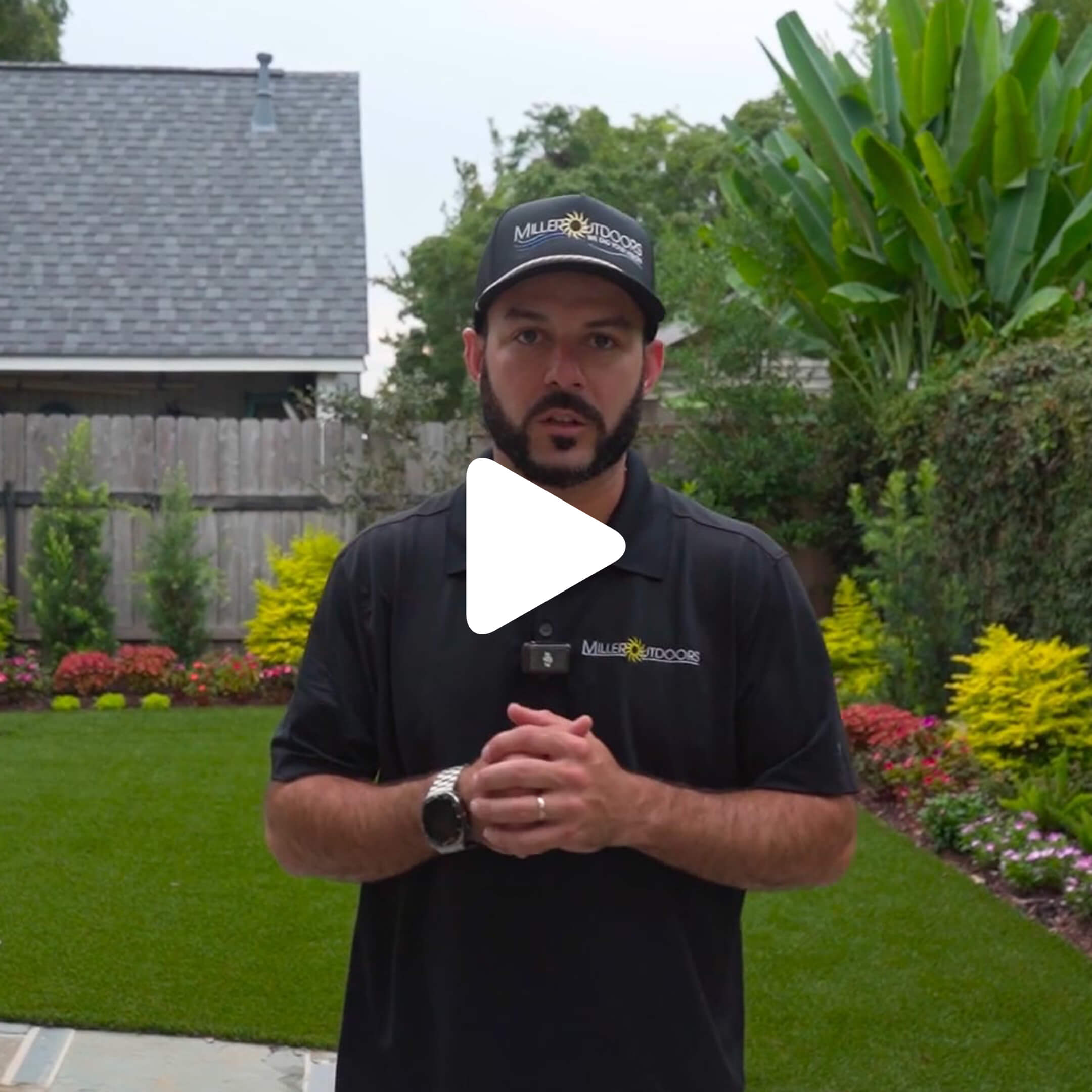 A man wearing a black polo shirt and cap with "MILLER OUTDOORS" stands on a well-manicured lawn, perfect for sharing gardening tips on a blog, with trimmed hedges, vibrant flowers, and a wooden fence in the background. A play button overlays the image.