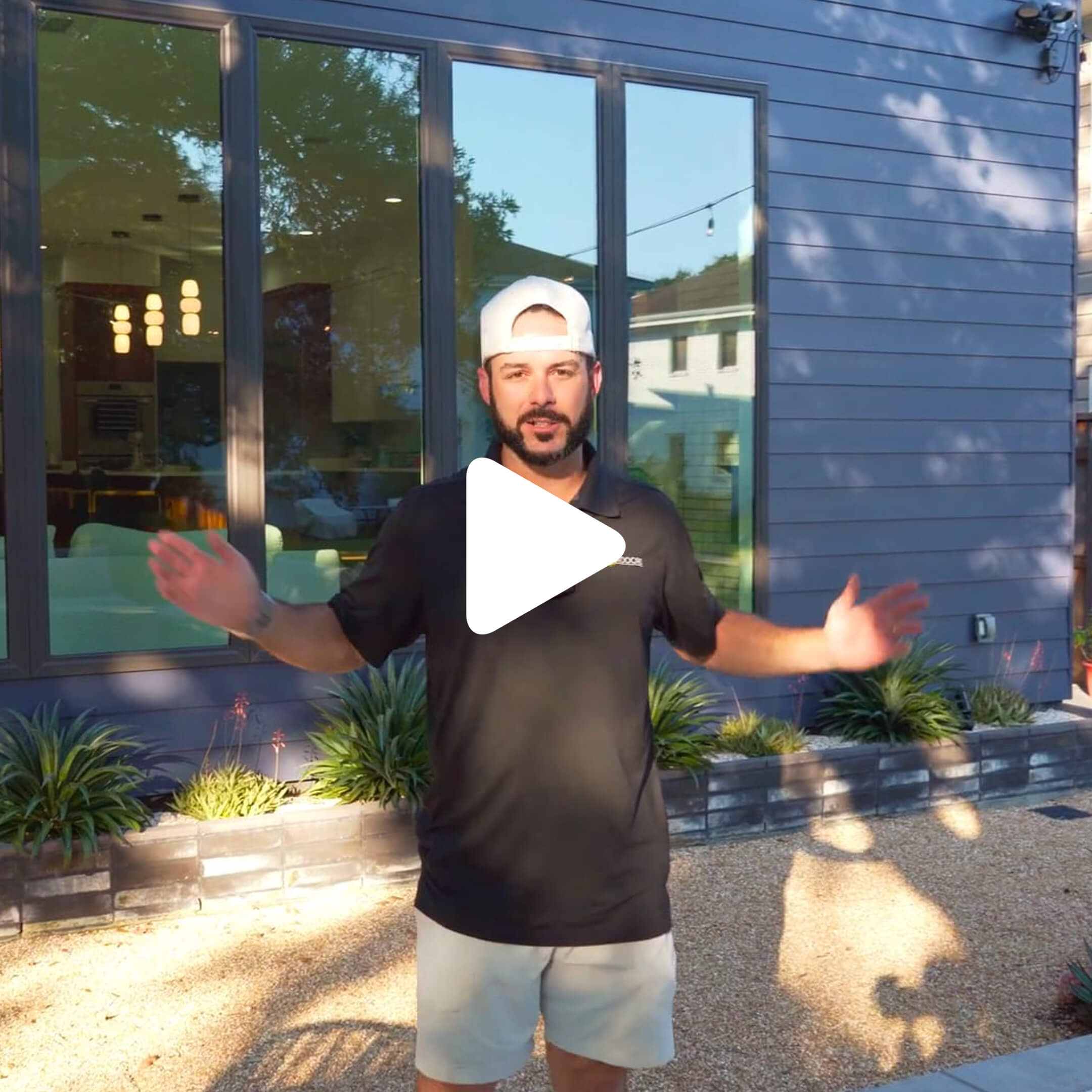 A man wearing a white cap, black shirt, and light shorts stands outside in front of a modern house with large windows, surrounded by landscaping and gravel—perfect for a blog feature on contemporary architecture. His arms are spread apart.