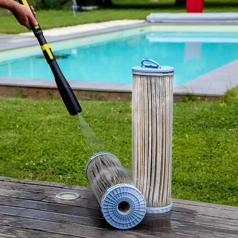 A person sprays water from a pressure washer onto two cylindrical pool filters, one standing upright and the other lying on its side, on a wooden deck near an outdoor pool.