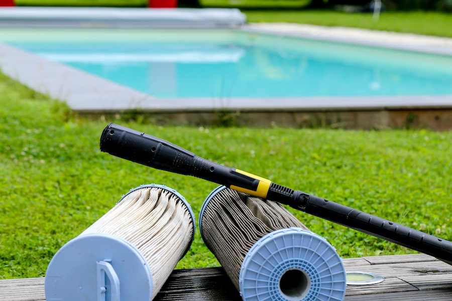 Two pool filters, one dirty and one clean, are placed side by side on a wooden surface outdoors, with a pressure washer wand resting on them and a swimming pool and green grass visible in the background.