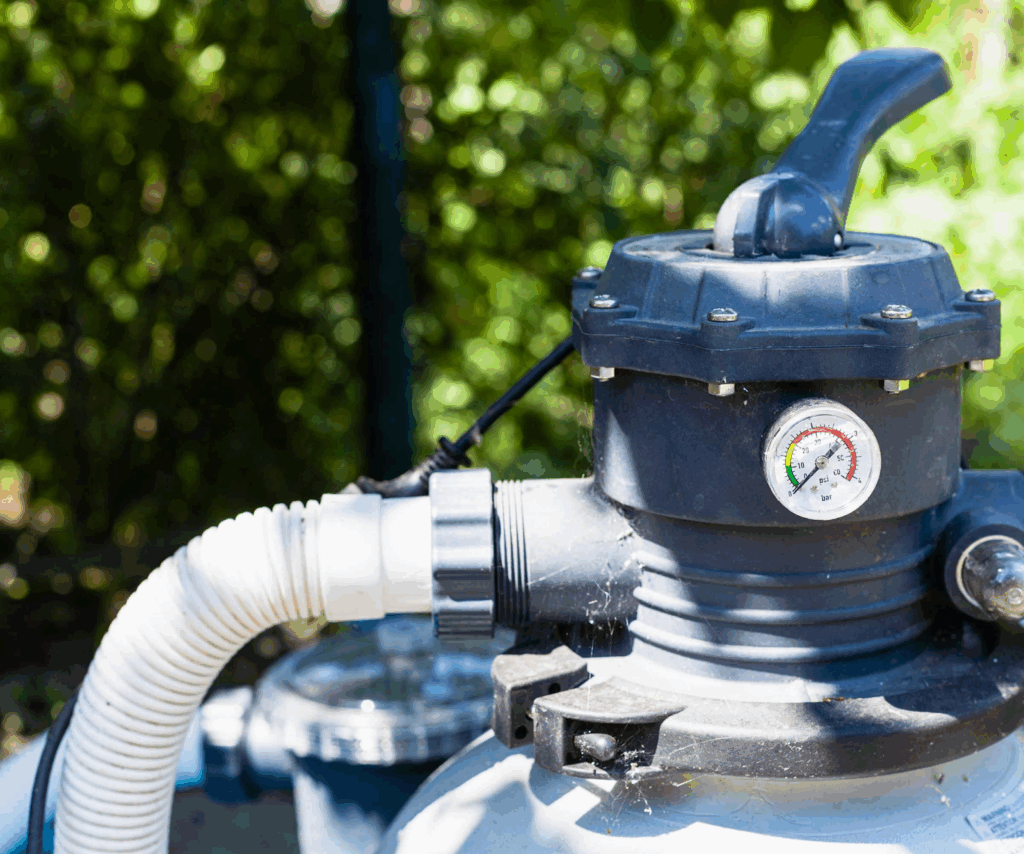 A close-up of a pool filter pump with a pressure gauge showing the needle in the yellow caution zone. A white hose is attached, and trees are visible in the blurred background.