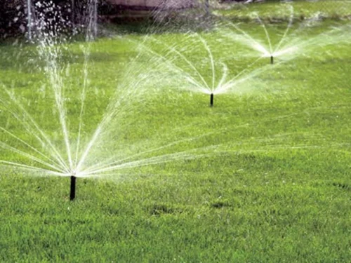 Three sprinklers are evenly spaced on a green lawn, spraying water in wide arcs to irrigate the grass. The scene shows healthy, well-maintained grass with visible water droplets in the air.