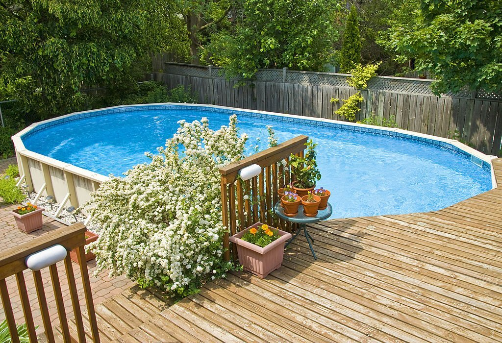 An above-ground vinyl pool is surrounded by a wooden deck with potted plants and a flowering shrub. Homeowners will appreciate the inviting space, while a wooden fence and green trees line the background.