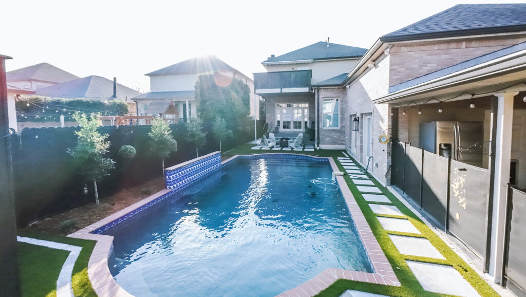 A long, narrow backyard pool bordered by grass and stone pavers is shown at the Ghafoor Residence between two-story houses. Outdoor seating awaits at the far end, with a covered patio and grill on the right side.