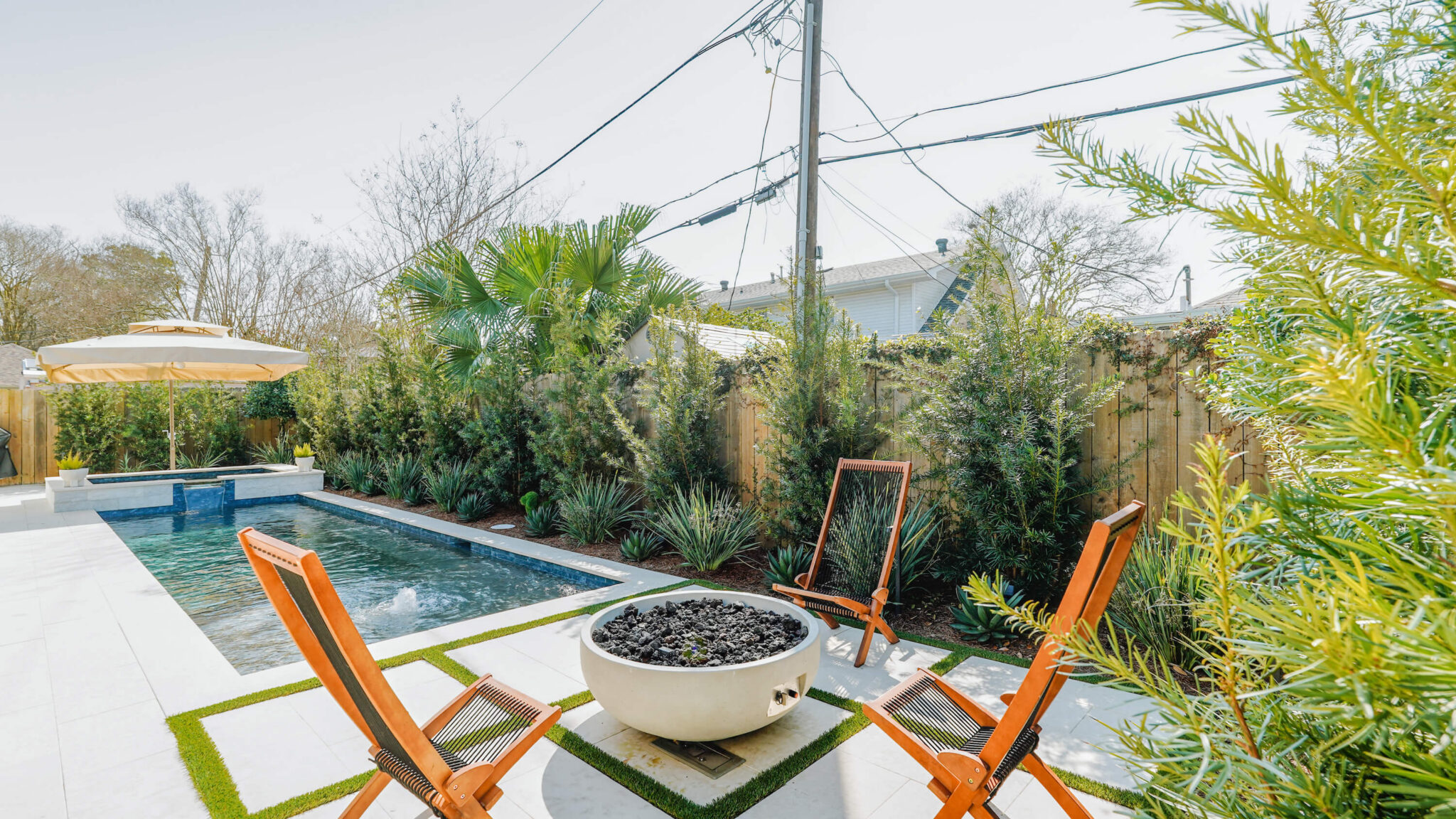 A backyard at the Versailles Residence features a rectangular swimming pool, a fire pit encircled by wooden chairs, and tall green plants along a wooden fence. Power lines and the house are visible in the background.
