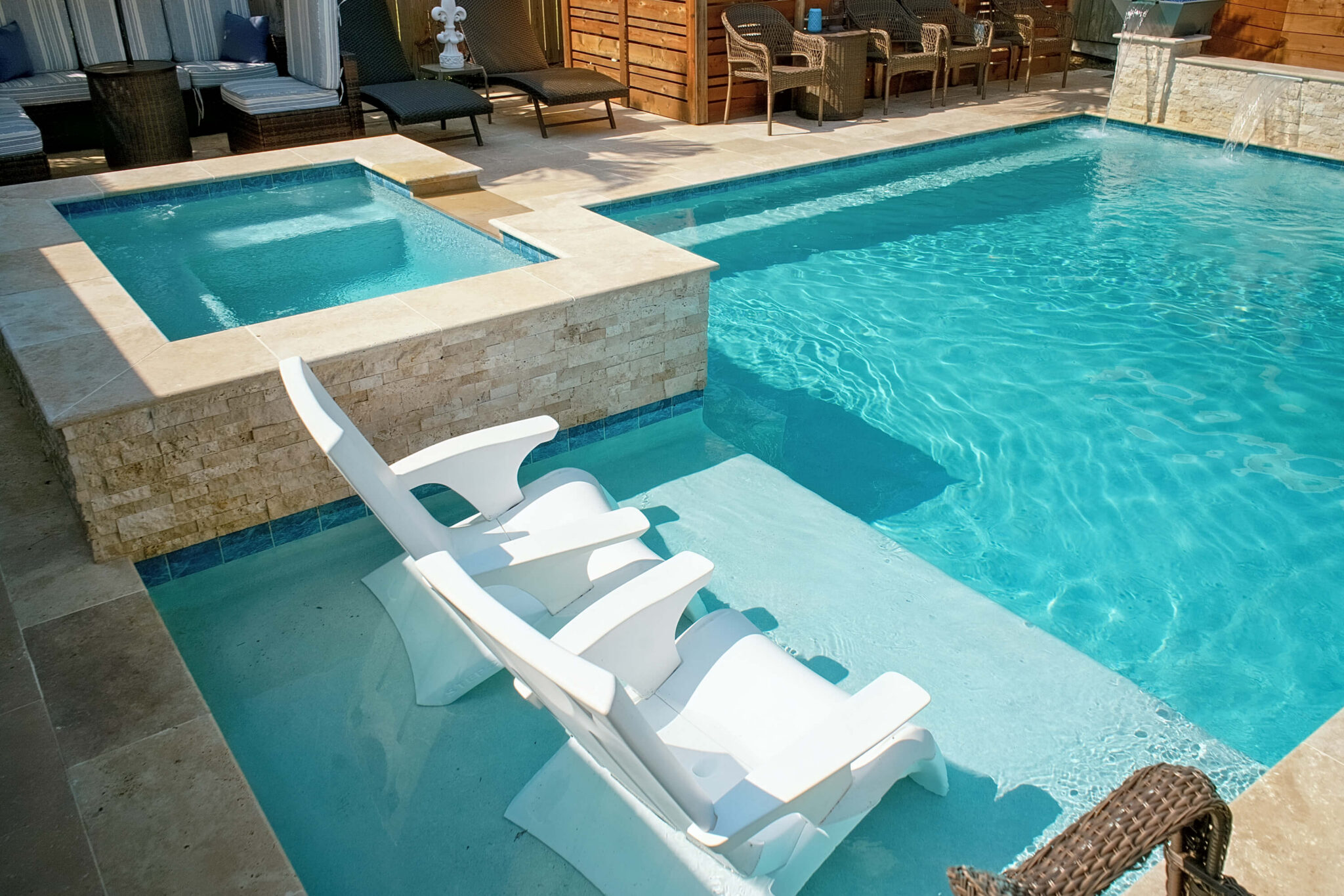 Two white lounge chairs rest on a shallow ledge in the Murphy Residence’s backyard pool, which features a built-in hot tub and is surrounded by stone tiles. Patio furniture can be seen in the background.