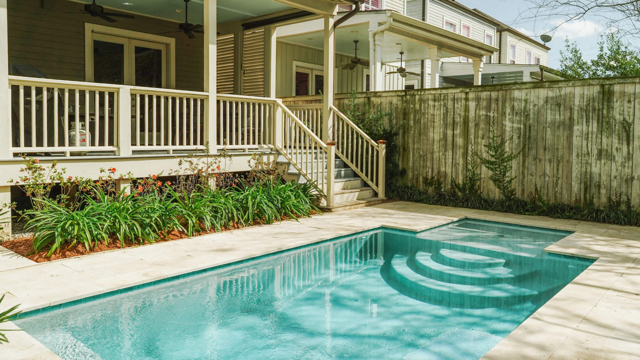 At the Rau Residence, a small rectangular backyard swimming pool with a built-in spa sits amid a stone patio. A wooden fence and green plants line one side, and a covered porch with stairs leads down from the house.