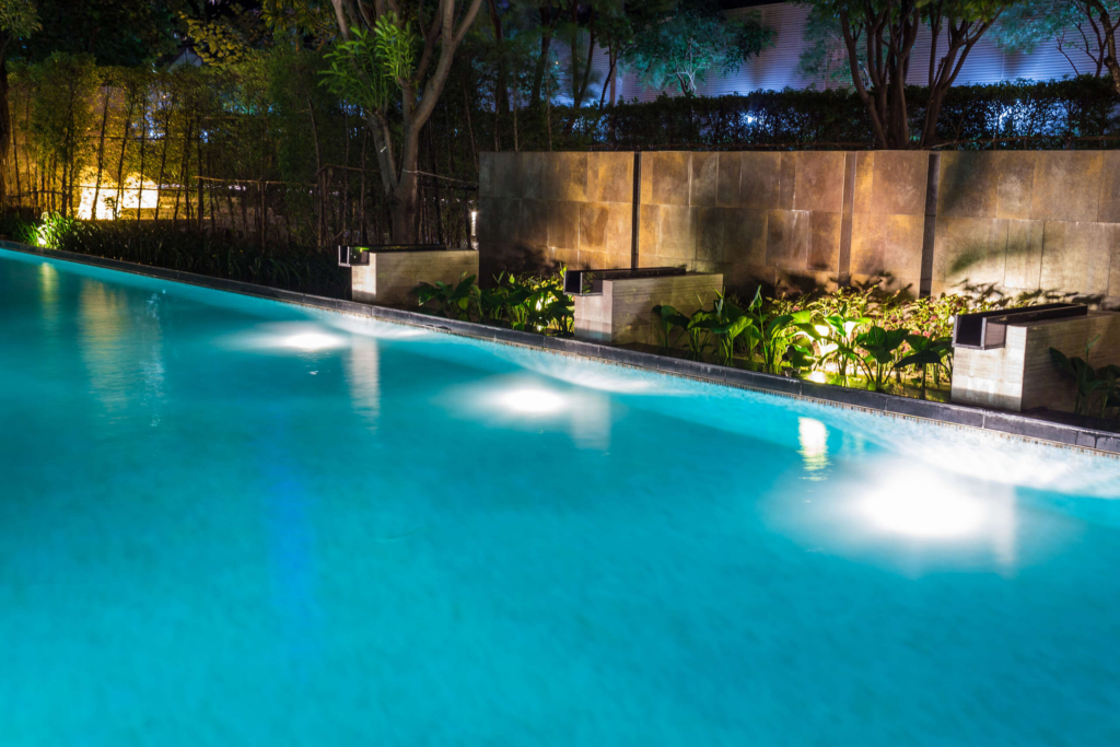 A high tech swimming pool at night features underwater lights illuminating the blue water. The pool is bordered by plants and a stone wall, with trees and additional lighting in the background.